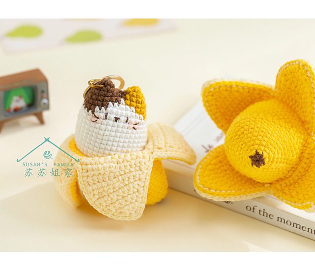 Una Figura Tejida a Crochet De Un Gato Blanco Con Un Traje Hecho Con Cáscaras De Plátano Se Sienta Junto a Un Narciso Amarillo Tejido a Crochet Sobre Una Superficie De Color Blanquecino En El Fondo Hay Un Pequeño Televisor Verde Que Muestra Una Caricatura Y La Portada De Un Libro La Escena Es Acogedora Y Caprichosa