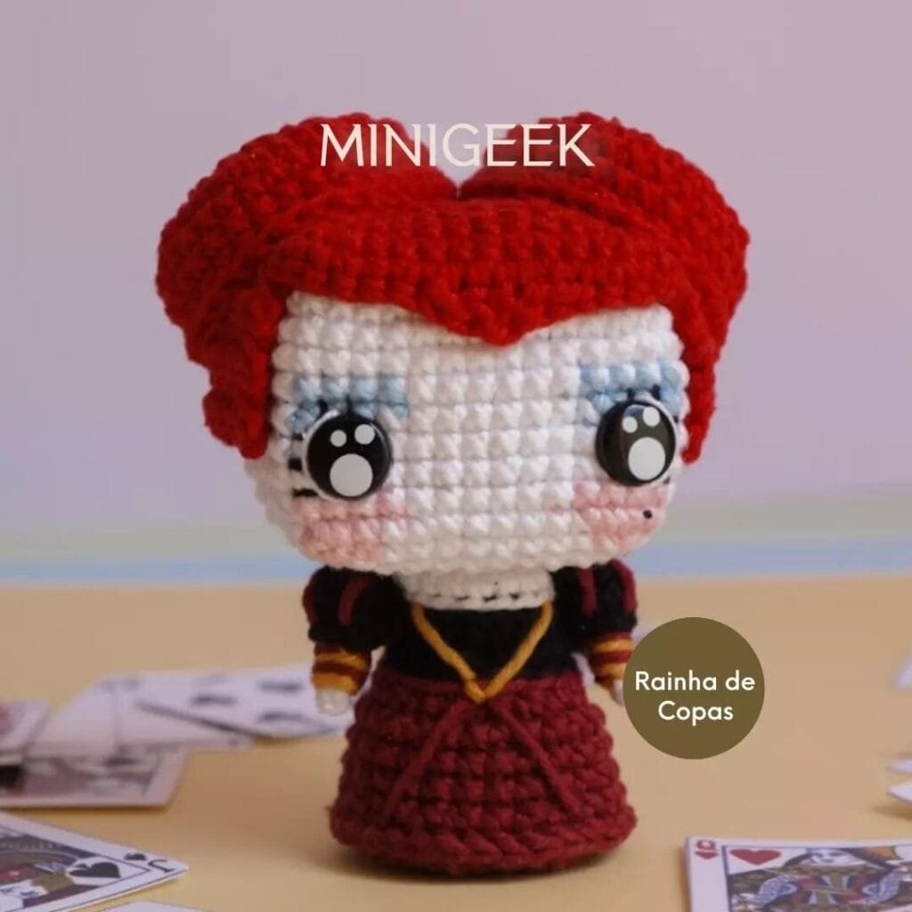 Una linda muñeca de crochet Reina de Corazones con rasgos exagerados, asemejándose a la Reina de Corazones. La muñeca tiene ojos grandes y redondos, cabello rojo peinado en forma de corazón y está vestida con un traje rojo y negro. Los naipes están esparcidos por la base. El texto "MINIGEEK" y "Rainha de Copas" está presente.