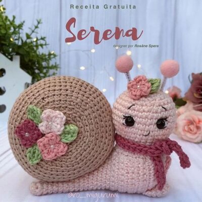 Serena, un caracol tejido a crochet diseñado por Rosáne Spera, se muestra con una concha beige decorada con flores rosas, blancas y verdes. El caracol tiene un delicado lazo rosa alrededor del cuello y está sobre un fondo con flores y vegetación.