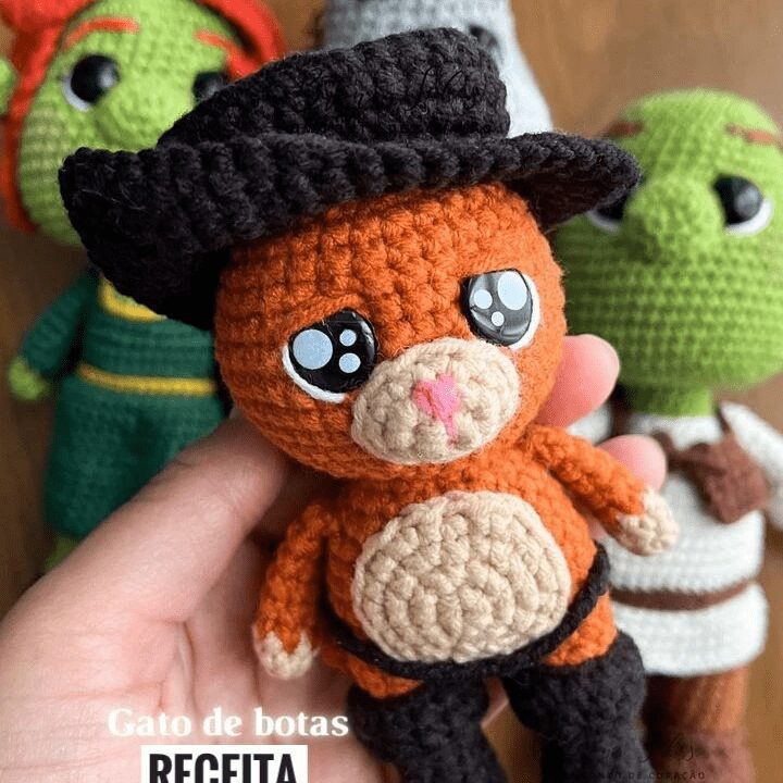 Una Mano Sostiene Un Muñeco Amigurumi Gato Con Botas De Ganchillo Con Ojos Grandes Un Sombrero Negro Y Botas Al Fondo Hay Otros Muñecos Tejidos a Crochet Entre Ellos Un Ogro Y Otro Personaje El Texto En La Parte Inferior Dice gato De Botas Receita