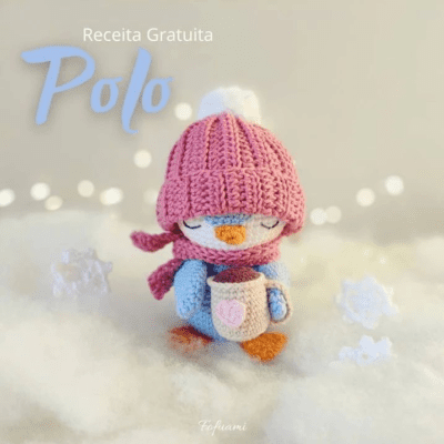 Un pingüino de crochet con un gorro y una bufanda de punto rosa sostiene una taza con un diseño de corazón, de pie sobre una superficie blanca y esponjosa. El texto lee "Receita Gratuita Polo". Las decoraciones de copos de nieve y la iluminación tenue crean un ambiente acogedor de invierno, perfecto para quienes buscan una solución gratuita de amigurumi.