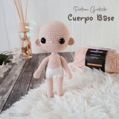 Una muñeca amigurumi tejida a crochet en tonos nude y con grandes ojos negros se encuentra sobre una alfombra blanca y esponjosa. Detrás hay ovillos de lana y agujas de tejer. Las palabras "Patrón Gratuito Cuerpo Base" están escritas en la parte superior derecha y el identificador de Instagram "@rukigurumis" es visible en la parte inferior izquierda.