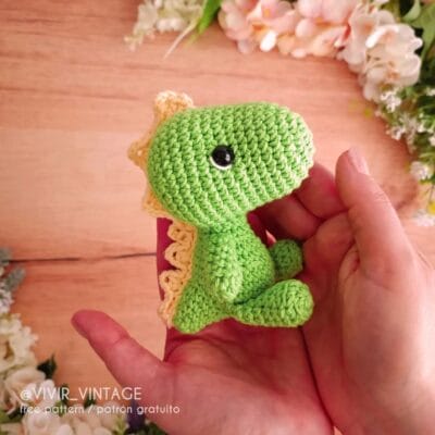 Una persona sostiene un pequeño dinosaurio verde tejido a crochet con púas de color crema a lo largo de la espalda. El fondo incluye una superficie de madera clara y flores blancas. El texto en la imagen dice "@VIVIR_VINTAGE free pattern / patrón gratuito".