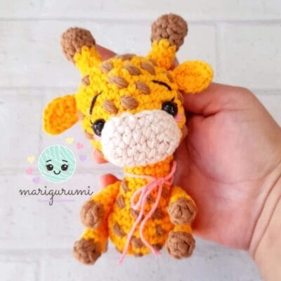 Una mano sostiene una pequeña jirafa tejida a crochet con un cuerpo de color amarillo brillante y naranja, manchas marrones y pezuñas marrones. La jirafa tiene ojos negros, un lazo rosa alrededor del cuello y lindas facciones faciales. El fondo es una pared de ladrillos blancos. En la imagen se lee "marigurumi".