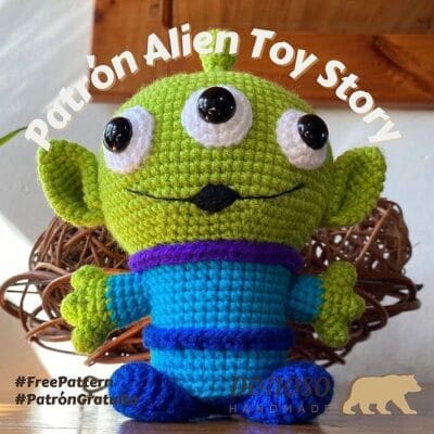 Peluche de crochet del personaje extraterrestre verde de tres ojos de Toy Story. El extraterrestre tiene un atuendo azul y morado y está colocado sobre un fondo con una decoración de mimbre. El texto en la imagen dice "Patrón Alien Toy Story", "#FreePattern #PatrónGratis" y "Dado80 Handmade".