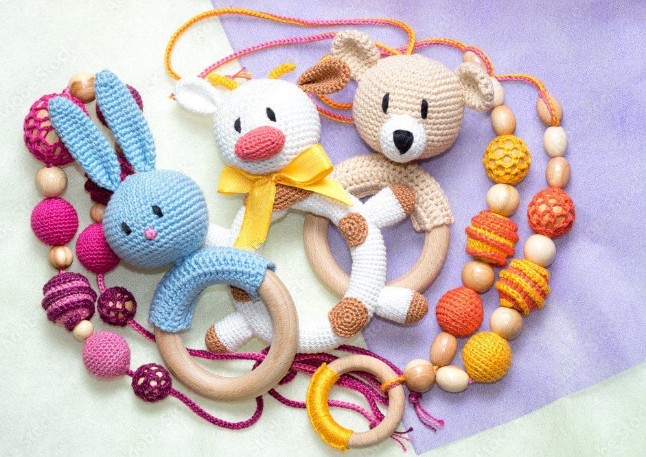 Una Colección De Coloridos Juguetes Para Bebés Amigurumi De Ganchillo Se Encuentra Sobre Un Fondo Pastel El Conjunto Incluye Un Conejo Azul Un Pato Blanco Y Un Oso Color Canela Cada Uno De Ellos Sujeto a Anillos De Madera Además Hay Varios Collares De Cuentas En Tonos Vibrantes De Rosa Amarillo Y Naranja