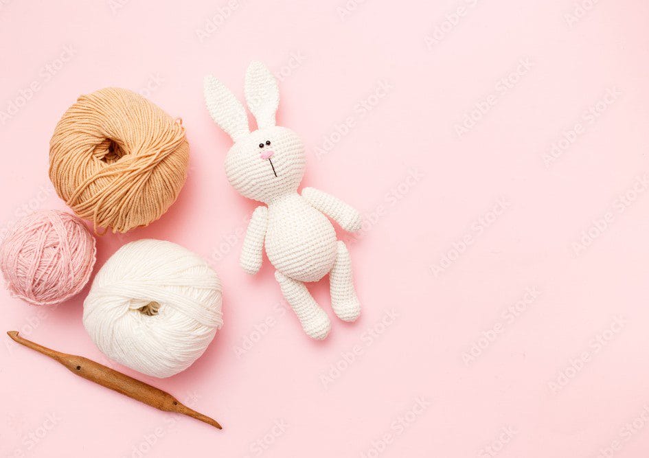 Un Conejito De Ganchillo Yace Sobre Un Fondo Rosa Junto a Ovillos De Hilo En Beige Melocotón Y Blanco Se Coloca Un Ganchillo De Madera Debajo Del Hilo Que Muestra Los Suministros Perfectos Para Crear Adorables Muñecos De Crochet Amigurumi