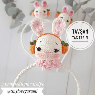 Una diadema tejida a crochet con un lindo diseño de conejito, con hilo blanco y detalles en naranja para las orejas y las orejeras. El fondo tiene un tono grisáceo suave. El texto en turco en la imagen dice "Tavşan Taç Tarifi". Los perfiles en redes sociales son @pembedugmeatolye y @tinylovegurumi.