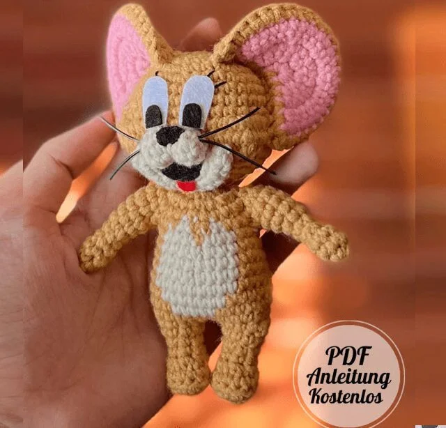 Una Mano Sostiene Un Peluche Tejido a Crochet Que Se Asemeja a Un Ratón Con Orejas Grandes Ojos Muy Abiertos Y Una Expresión Feliz El Texto pdf Anleitung Kostenlos Aparece En Una Placa Circular En La Esquina Inferior Derecha