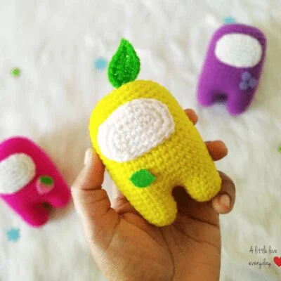 Mano que sostiene a un compañero de tripulación amarillo tejido a crochet del juego "Among Us" con una hoja verde en la cabeza. Al fondo, hay otros dos compañeros de tripulación tejidos a crochet, uno rosa y otro morado, sobre una superficie blanca. El texto en la esquina dice: "Un poco de amor todos los días ❤️".