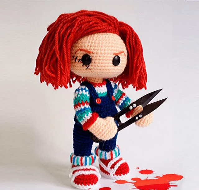 Un Muñeco De Ganchillo Con Pelo Rojo Que Recuerda a Chucky Vestido Con Un Traje Colorido Que Incluye Una Camisa a Rayas Un Mono Azul Y Zapatos Rojos Sostiene Un Par De Tijeras La Muñeca Se Encuentra Sobre Una Superficie Blanca Con Manchas Rojas Que Parecen Manchas De Sangre