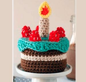 Un pastel de crochet con capas marrones, blancas y azules está cubierto con formas de fresas rojas y una vela de crochet con una llama amarilla y roja. La pieza decorativa se coloca sobre un soporte para tartas blanco.