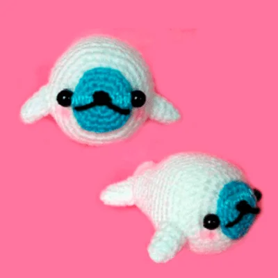 Dos peluches de ganchillo que se asemejan a animales marinos blancos y regordetes con caras azules y ojos negros. Tienen pequeñas aletas a los lados y en la espalda. El fondo es de color rosa intenso.