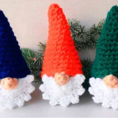 Tres gnomos tejidos a crochet con esponjosas barbas blancas están uno al lado del otro. El gnomo de la izquierda tiene un sombrero azul, el del medio tiene un sombrero naranja y el de la derecha tiene un sombrero verde. Están colocadas sobre un fondo claro con una ramita de vegetación detrás de ellas.