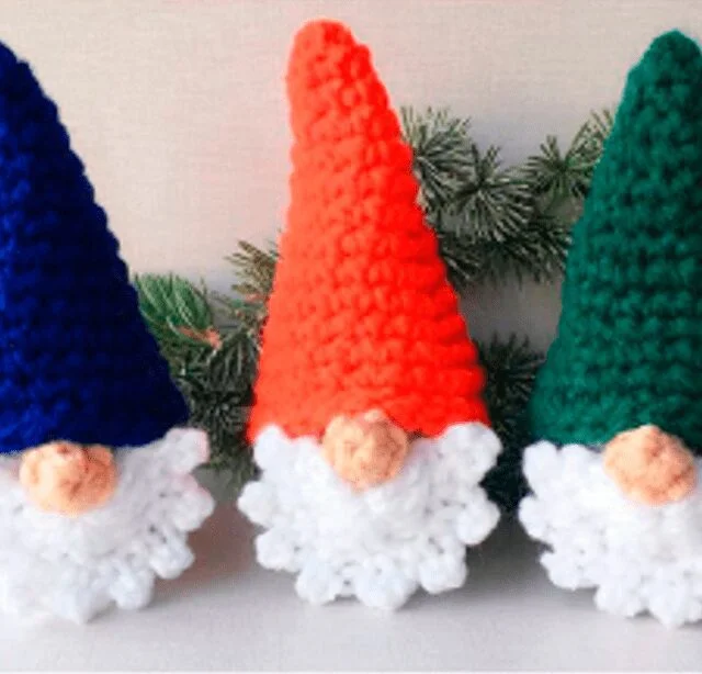 Tres Gnomos Tejidos a Crochet Con Esponjosas Barbas Blancas Están Uno Al Lado Del Otro El Gnomo De La Izquierda Tiene Un Sombrero Azul El Del Medio Tiene Un Sombrero Naranja Y El De La Derecha Tiene Un Sombrero Verde Están Colocadas Sobre Un Fondo Claro Con Una Ramita De Vegetación Detrás De Ellas