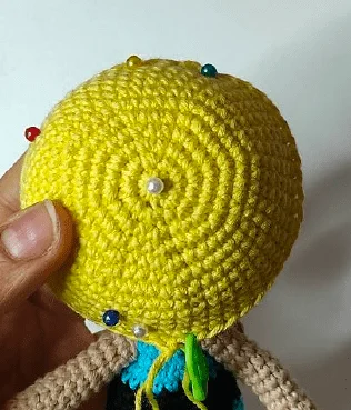 Una Persona Sostiene Un Objeto Redondo Amarillo Tejido a Crochet Posiblemente Parte De Una Muñeca De Chicas Superpoderosas Con Pequeños Alfileres De Colores Insertados En él El Pulgar Y El índice De La Persona Son Visibles Sosteniendo El Objeto El Fondo Es Sencillo Y Desenfocado