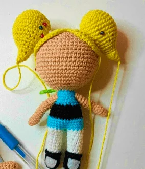 Se Está Desarrollando Una Muñeca De Crochet Inspirada En Las Chicas Superpoderosas Con Una Cabeza De Color Marrón Claro Un Peinado Amarillo Sujeto Con Alfileres Y Un Cuerpo Parcialmente Terminado Con Un Traje Azul Y Negro Cerca Se Ven Hilos Sueltos Y Un Ganchillo