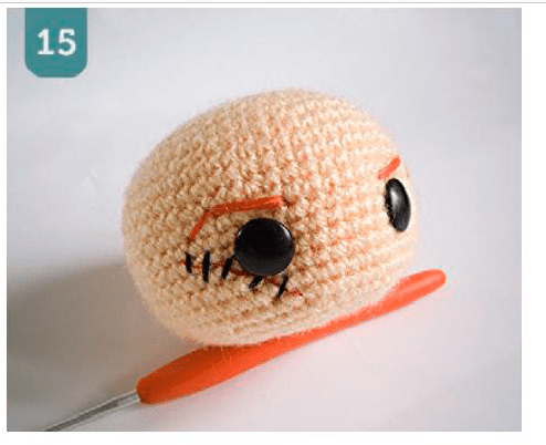 Una Cabeza Pequeña Y Redonda De Crochet Con Grandes Ojos Negros Y Cejas Anaranjadas Emite Una Vibra Juguetona Que Recuerda a Chucky Al Lado Hay Un Ganchillo Naranja Los Puntos De Crochet Son Visibles Y Un Número 15 En Un Cuadrado Azul Se Encuentra En La Esquina Superior Izquierda