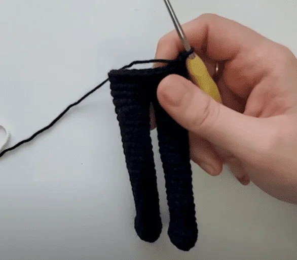 Una Mano Que Sostiene Un Ganchillo Está Trabajando En Un Pequeño Proyecto De Crochet El Proyecto Está Hecho De Hilo Oscuro Y Parece Formar Dos Formas Estrechas Y Alargadas Que Se Asemejan a Patas El Ganchillo Tiene Un Mango Amarillo