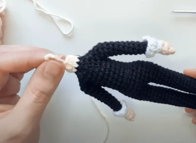 Primer Plano De Manos Sosteniendo Un Muñeco De Ganchillo Con Traje Negro Y Puños Blancos El Muñeco Está En Proceso De Confección Con Hilo Visible Y Una Aguja De Crochet Al Fondo