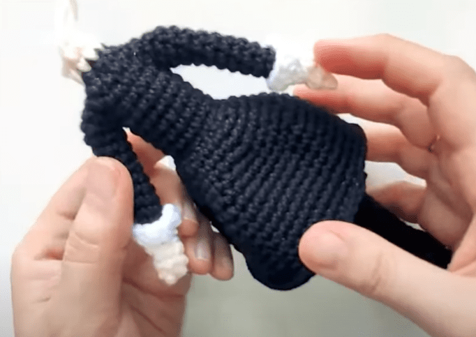 Una Persona Sostiene Una Pequeña Muñeca De Crochet Hecha a Mano Con Un Vestido Negro Cuello Blanco Y Puños Blancos La Muñeca No Tiene Cabeza Y Los Dedos De La Persona Acunan Suavemente Su Cuerpo Contra Un Fondo Liso