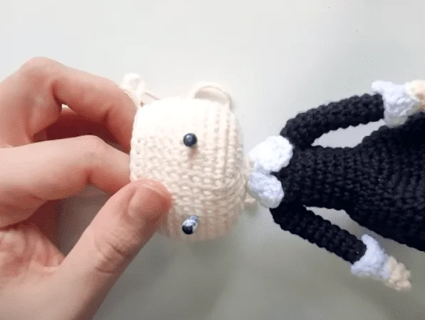 Un Primer Plano De Manos Armando Una Muñeca De Crochet Una Mano Sostiene La Cabeza Color Crema Con Ojos De Botón Negros Mientras Que La Otra Guía El Accesorio Al Cuerpo Blanco Y Negro De La Muñeca La Muñeca Parece Llevar Un Traje Formal Posiblemente Un Esmoquin