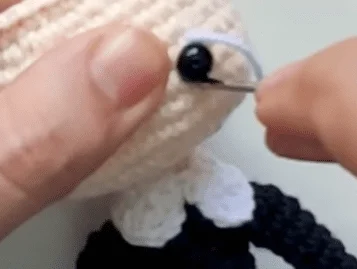 Primer Plano De Manos Cosiendo Un Ojo De Cuentas Negras En Una Muñeca Tejida O De Ganchillo La Muñeca Es Principalmente De Color Crema Con Algunos Detalles De Hilo En Blanco Y Negro Visibles Una Aguja Está En El Proceso De Colocar La Cuenta