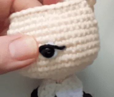 Un Primer Plano De Una Persona Sosteniendo Una Muñeca De Crochet Hecha a Mano La Muñeca Tiene Hilo De Color Claro Y Un Pequeño Botón Negro Con Una Pestaña De Fieltro O Hilo Negro La Mano Izquierda De La Persona Ajusta O Sostiene El Muñeco Cerca De Su Ojo