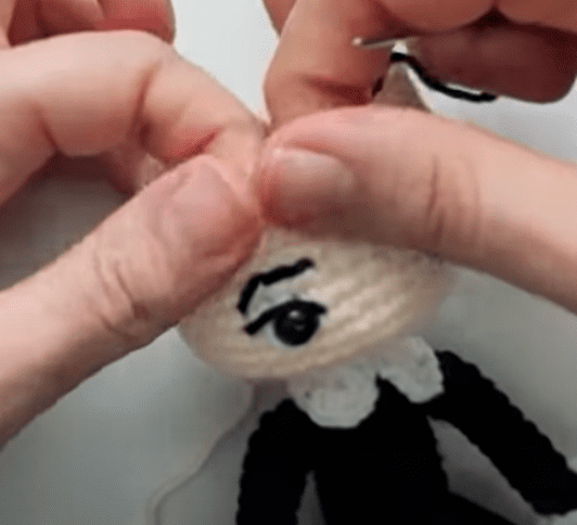 Vista De Primer Plano De Las Manos Cosiendo La Parte Superior De Una Cabeza De Muñeca De Ganchillo Que Presenta Un Cuello Blanco Ropa Negra Y Un Solo Ojo Bordado Con Una Ceja