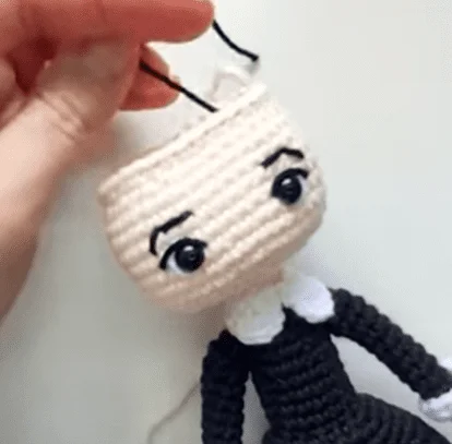 Una Mano Sostiene Una Muñeca De Crochet Parcialmente Terminada Por La Parte Superior De Su Cabeza La Muñeca Tiene Ojos Negros Grandes Y Redondos Y Una Cara Sencilla Con Cejas Negras Está Vestido Con Un Traje Negro Con Cuello Blanco