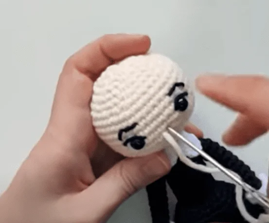 Dos Manos Sostienen Una Cabeza De Muñeca De Crochet Con Ojos Y Cejas Bordados Una De Las Manos Está Usando Un Ganchillo Para Agregar Detalles a La Cabeza La Muñeca De Crochet Sin Terminar Es Blanca Con Detalles De Hilo Negro