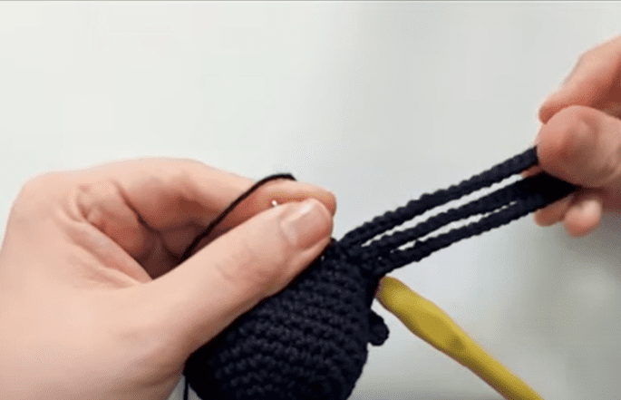 Un Primer Plano De Las Manos De Alguien Sosteniendo Una Pieza De Crochet De Color Oscuro En Proceso Una Mano Sostiene Los Bucles De Hilo Mientras Que La Otra Maniobra Un Ganchillo Amarillo a Través De Los Puntos El Fondo Es Liso Y Borroso