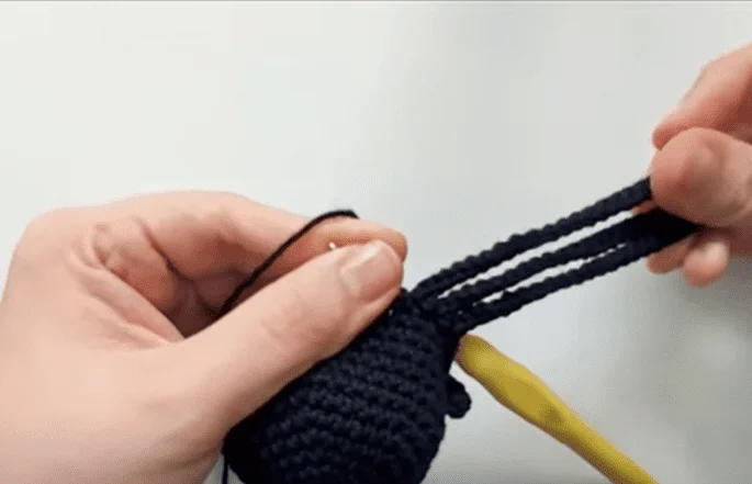 Un Primer Plano De Las Manos De Alguien Sosteniendo Una Pieza De Crochet De Color Oscuro En Proceso Una Mano Sostiene Los Bucles De Hilo Mientras Que La Otra Maniobra Un Ganchillo Amarillo a Través De Los Puntos El Fondo Es Liso Y Borroso