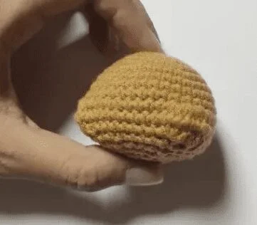 Una Mano Sostiene Un Objeto Pequeño Y Redondo De Crochet Que Se Asemeja a Un Gorro De Hongo Hecho De Hilo Marrón Y Tostado El Fondo Es Completamente Blanco