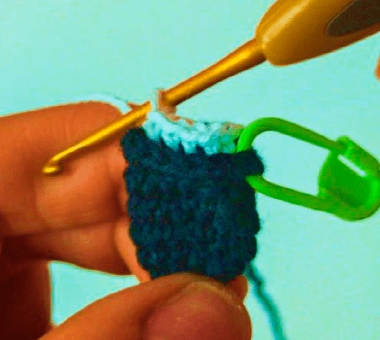 Primer Plano De Alguien Tejiendo Con Hilo Azul Oscuro La Persona Sostiene Un Marcador De Puntadas Verde Y Un Ganchillo Trabajando En Una Pequeña Pieza De Crochet Con Un Borde Superior Azul Claro Sobre Un Fondo Azul Claro Es Tan Complejo Como Cualquier Manualidad De Chicas Superpoderosas