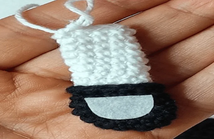 Un Pequeño Adorno Hecho a Mano En Crochet Con Forma De Figura Con Cuerpo Blanco Base Negra Parecida a Una Barba Y Un Lazo En La Parte Superior El Adorno Que Recuerda Al Amuleto De Chicas Superpoderosas Se Sostiene En La Mano De Una Persona Contra La Palma