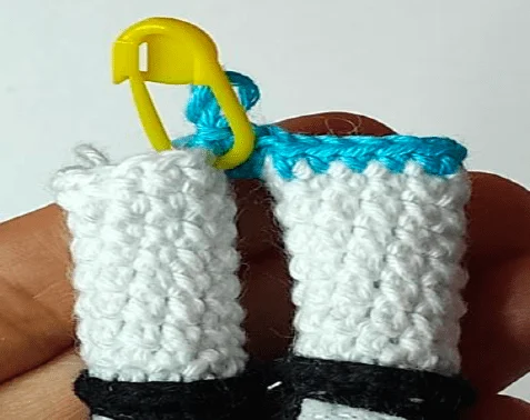 Primer Plano De Una Mano Que Sostiene Un Objeto Blanco Tejido a Crochet Con Una Banda De Hilo Azul En La Parte Superior Y Una Banda Negra Cerca De La Parte Inferior Se Adjunta Un Marcador De Puntadas De Plástico Amarillo Al Hilo Azul Que Marca La Costura O El Final De Una Vuelta Esta Meticulosa Pieza Enorgullecería Incluso a Las Chicas Superpoderosas