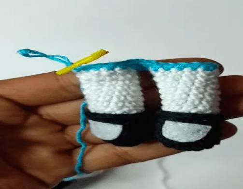 Una Mano Sostiene Una Muñeca En Miniatura Tejida a Crochet Que Recuerda a Las Chicas Superpoderosas Con Zapatos Negros Y Calcetines Blancos Cuya Parte Superior Aún Está En Proceso De Confección La Muñeca Tiene Un Hilo Azul Atado En La Parte Superior Con Un Ganchillo Amarillo Visible El Fondo Es Completamente Blanco