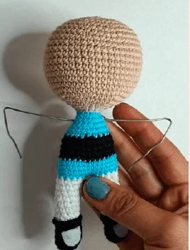 Una Muñeca De Crochet De Chicas Superpoderosas En Proceso Es Sostenida Por Una Mano Con Uñas Pintadas De Azul La Muñeca Tiene Una Cabeza Sin Terminar Hecha De Hilo Beige Un Cuerpo Azul Claro Con Una Raya Negra Piernas Blancas Zapatos Negros Y Brazos De Alambre Expuestos Sin Cubierta De Hilo