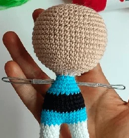 Una Mano Que Sostiene Una Muñeca De Crochet En Proceso Se Asemeja a Una De Las Chicas Superpoderosas La Muñeca Tiene Una Cabeza Grande Y Redonda Un Torso a Rayas Azules Y Negras Y Piernas Blancas Los Brazos Son Simplemente Marcos De Alambre Y La Cara Queda Sin Terminar