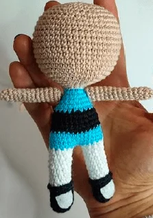 Un Primer Plano De Una Muñeca De Crochet Sostenida En La Mano De Una Persona La Muñeca Que Se Asemeja a Una De Las Chicas Superpoderosas Tiene La Cabeza Beige Brazos Extendidos Una Blusa a Rayas Azules Y Negras Pantalones Blancos Y Zapatos Negros Al Fondo Se Ve La Mano Que Sostiene La Muñeca