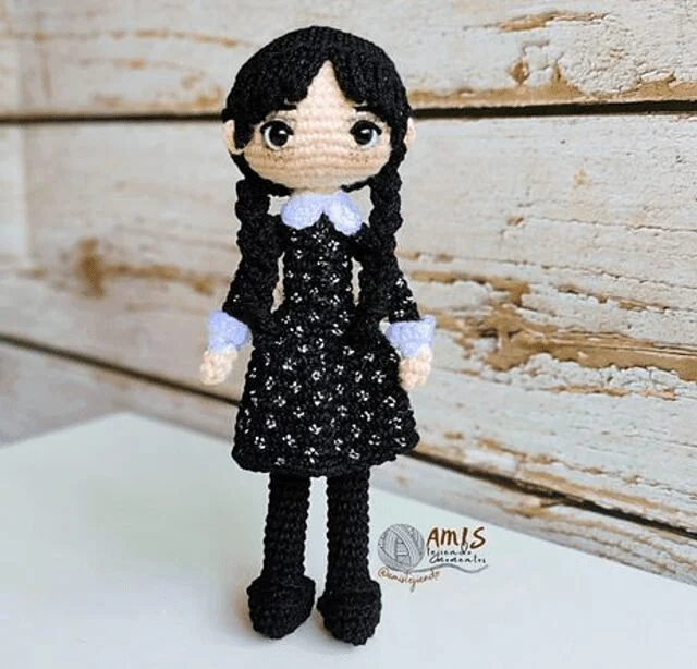 Una Muñeca De Ganchillo Se Alza Sobre Un Fondo De Madera La Muñeca Tiene Trenzas Negras Cuello Blanco Y Puños Blancos Sobre Un Vestido Negro La Figura Presenta Ojos Grandes Y Expresivos Y Detalles Faciales Minimalistas Que Muestran Una Artesanía Intrincada Y Atención Al Detalle