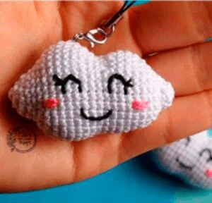 Una mano sostiene un pequeño dije de nube blanca tejido a crochet con una cara feliz, con ojos cerrados y una boca sonriente. El dije tiene marcas de rubor rosado en sus mejillas y está sujeto a un lazo de hilo negro para colgar. El fondo es borroso y azul.