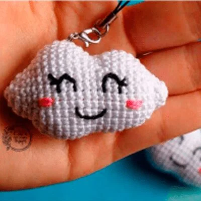 Una mano sostiene un pequeño dije de nube blanca tejido a crochet con una cara feliz, con ojos cerrados y una boca sonriente. El dije tiene marcas de rubor rosado en sus mejillas y está sujeto a un lazo de hilo negro para colgar. El fondo es borroso y azul.