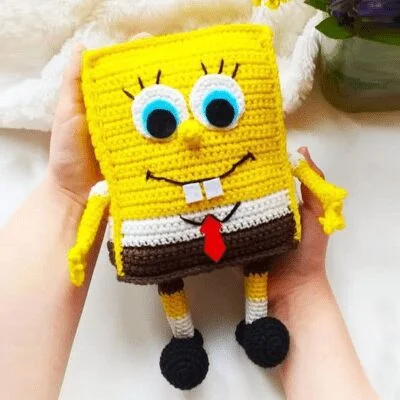 Un muñeco de crochet hecho a mano de Bob Esponja se sostiene suavemente con dos manos. El muñeco presenta detalles con las características icónicas de Bob Esponja, incluidos sus grandes ojos azules, camisa blanca, corbata roja, pantalones cuadrados marrones y zapatos negros.