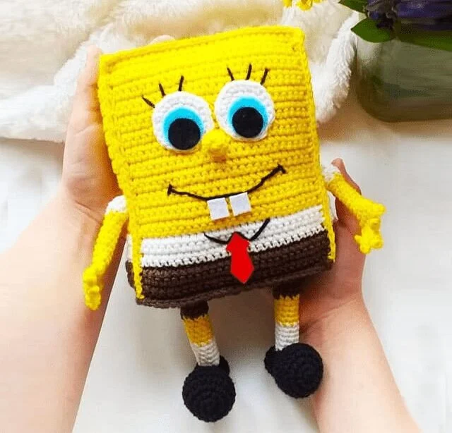 Un Muñeco De Crochet Hecho a Mano De Bob Esponja Se Sostiene Suavemente Con Dos Manos El Muñeco Presenta Detalles Con Las Características Icónicas De Bob Esponja Incluidos Sus Grandes Ojos Azules Camisa Blanca Corbata Roja Pantalones Cuadrados Marrones Y Zapatos Negros