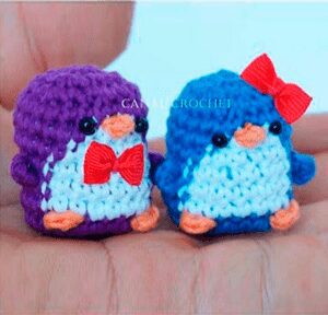 Primer plano de dos pequeñas figuras de pingüinos de ganchillo que descansan sobre la palma de una persona. El pingüino morado de la izquierda tiene una pajarita roja y el pingüino azul de la derecha tiene un lazo rojo en la cabeza. Ambos tienen pequeños ojos negros y picos y patas anaranjados.