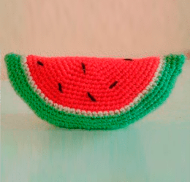 Una Prenda De Punto Diseñada Para Parecerse a Una Rodaja De Sandía La Pieza Presenta Una Corteza Verde Una Capa Interior Blanca Y Un Núcleo De Color Rojo Rosado Con Pequeños Puntos Negros Que Representan Semillas De Sandía El Artículo Está Apuntalado Para Mostrar Su Forma Semicircular