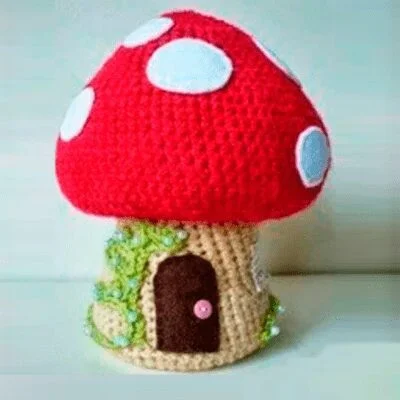 Una casa de setas tejida a mano con un gorro rojo y manchas blancas. El tallo beige tiene una pequeña puerta de color marrón oscuro adornada con un pomo de botón rosa y está decorado con enredaderas de hojas verdes. El artículo hecho a mano se apoya sobre un fondo liso.