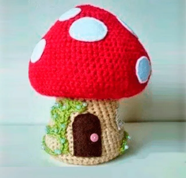 Una Casa De Setas Tejida a Mano Con Un Gorro Rojo Y Manchas Blancas El Tallo Beige Tiene Una Pequeña Puerta De Color Marrón Oscuro Adornada Con Un Pomo De Botón Rosa Y Está Decorado Con Enredaderas De Hojas Verdes El Artículo Hecho a Mano Se Apoya Sobre Un Fondo Liso